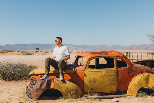 Young smiling man tourist in eyeglasses exploring the arid landscape with vintage abandoned cars in Namib desert, Southern Africa