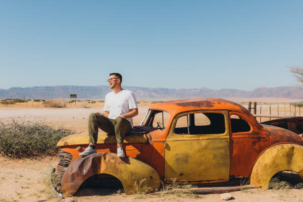 Happy male traveler sitting on the old abandoned car in the middle of the desert in Namibia stock photo