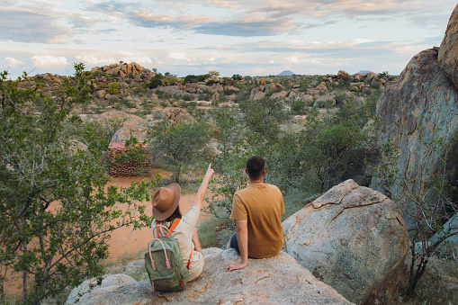 Young couple of woman in hat with backpack and man sitting on the top of the rock looking at the beautiful landscape and watching wild animals in Damaraland, Namibia, Southern Africa