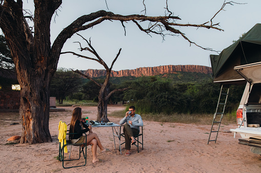 Young couple of female and male enjoying an outdoor dinner camping with their 4X4 camper truck in savannah under the trees with background view of the Watewrberg plateau, Southern Africa