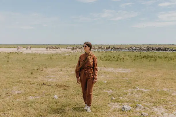 Young woman explorer in jumpsuit and sunglasses walking at the wild savannah looking at the group of wild animals in Southern Africa