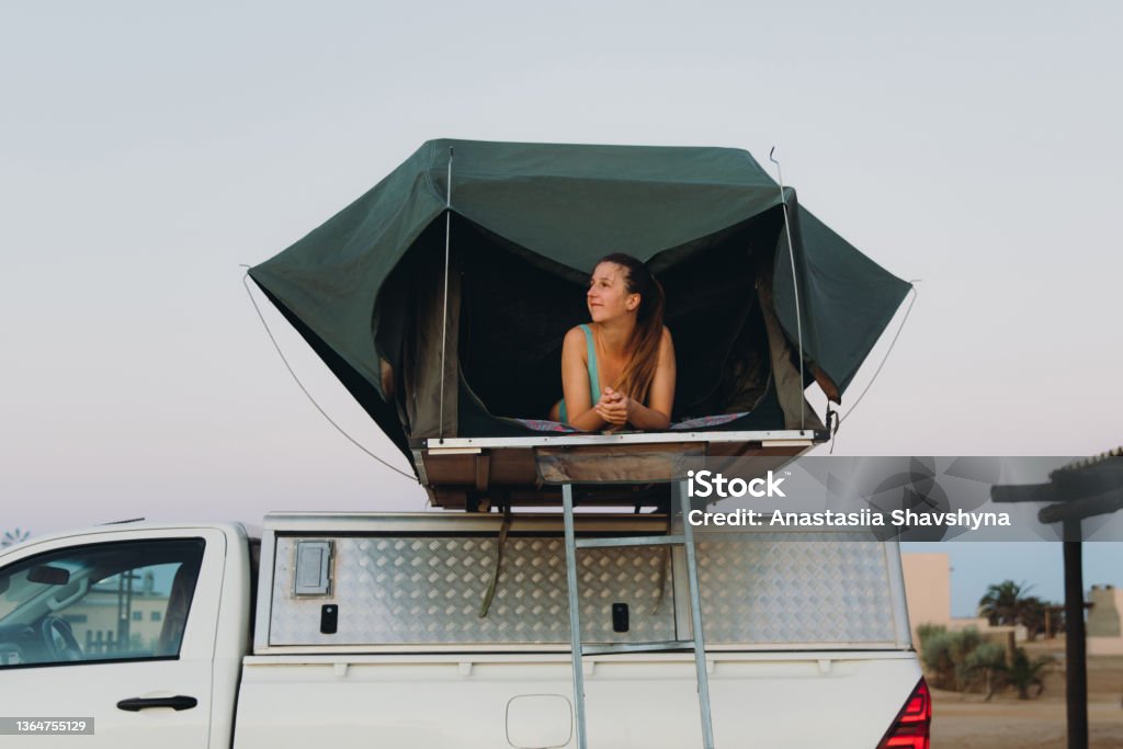 Happy female traveler relaxing in the camper truck by the beach in Namibia Smiling woman with long hair, in blue swimsuit lying in the raised roof tent above the 4X4 motor home admiring sunset at the Skeleton Coast, Southern Africa 20-29 Years Stock Photo