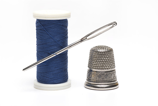 Close up of a blue roll with sewing thread of a sewing needle stuck in it and a silver thimble next to it on white background as a concept for sewing tailoring and repairing clothes