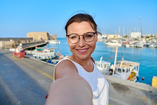 Middle-aged smiling woman taking selfie photo against the backdrop of historic fortress, sea bay. Tourism, travel, outdoor activities, Europe, Greece, Heraklion Koules Fortress