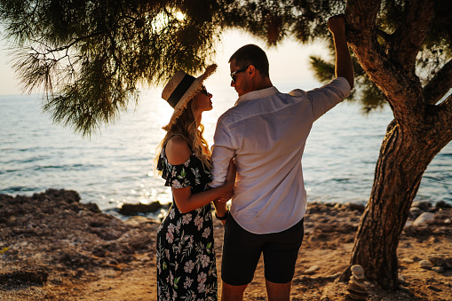 Loving young couple standing and hugging by the tree on coastline at summer sunset, side view