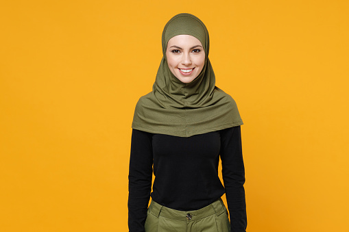 Smiling beautiful young arabian muslim woman wearing hijab black green clothes standing looking camera isolated on bright yellow color background, studio portrait. People religious lifestyle concept