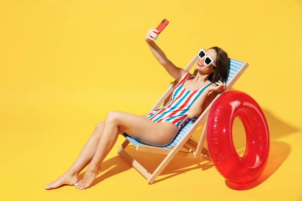 longueur complète du corps heureuse jeune femme porter maillot de bain bleu rouge s’asseoir sur une chaise en bois tenir utiliser téléphone portable isolé sur fond de couleur jaune vif studio été hôtel piscine repos de la mer concept de bronzage - deck chair photos et images de collection