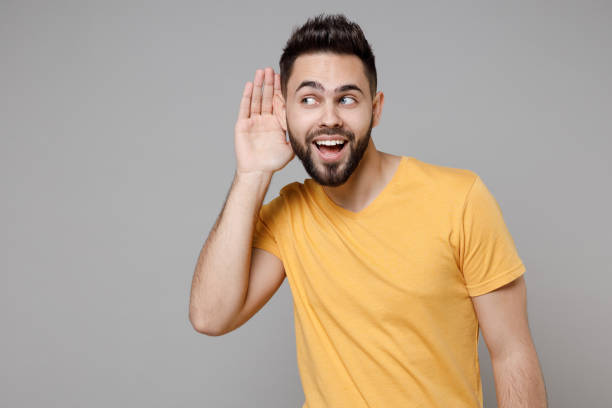 istock Young caucasian bearded smiling secret curious student man 20s wearing casual yellow basic t-shirt trying to hear you overhear listening intently isolated on grey color background studio portrait. 1364767684