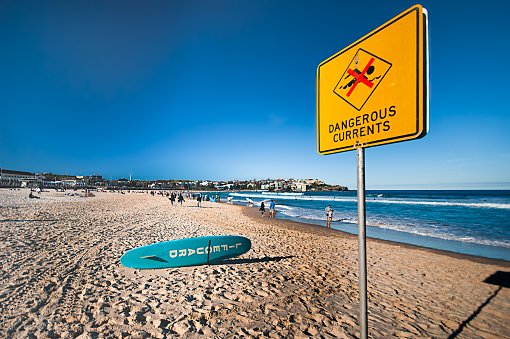 A bright yellow 'Dangerous Currents' warning sign is displayed on beach advising visitors not to swim, a blue lifeguard surfboard sits nearby