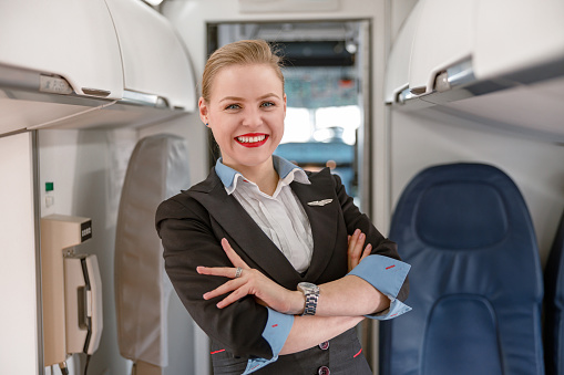 Joyful woman flight attendant keeping arms crossed and smiling while standing in aisle of aircraft salon