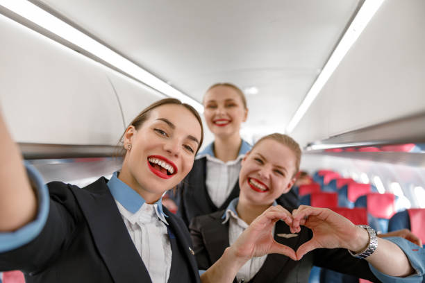 Cheerful flight attendants having fun in airplane Joyful woman stewardess doing heart symbol with hands and smiling while standing with colleagues in aircraft cabin air stewardess stock pictures, royalty-free photos & images