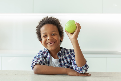 Happy small boy laughing and holding green apple. Healthy food and vitamins, smiling child on white kitchen background