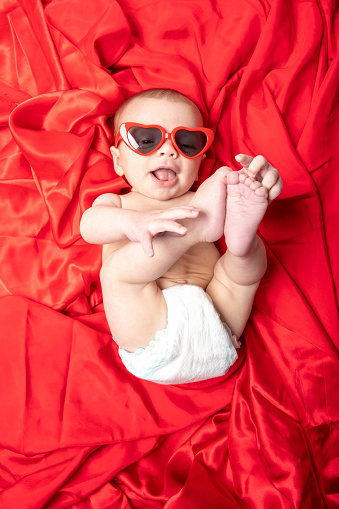 A 4 month old baby boy wearing heart shaped sunglasses on red satin.