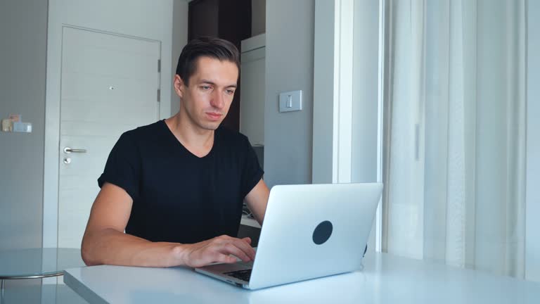 Young male freelancer using laptop processing images on his laptop and drinking coffee at home.