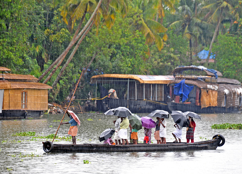 Men with umbrellas stand on a crowded punt  in the rain, Cochin, Kerala waterways, India. Kerala is a colourful Indian state of waterways and tropical rainforest geography on the southwestern Malabar Coast of the country owing its growth to the spice trade created by its Portuguese and British colonial occupiers that is now an attraction for tourists with its river boats and canals