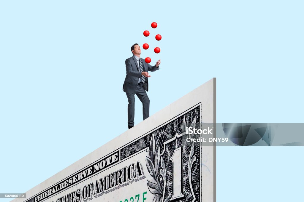Balancing  And Juggling Finances A man balances on the edge of a one dollar bill as he juggles several red balls isolated on a blue background. Inflation - Economics Stock Photo