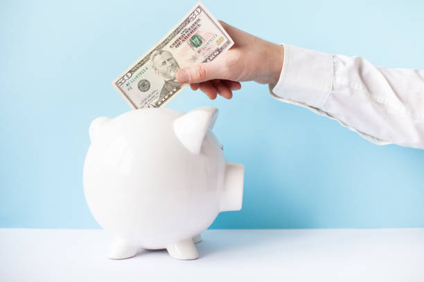 istock Mans hand puting 50 dollar banknote in  white piggy bank on a blue background. 1364807981