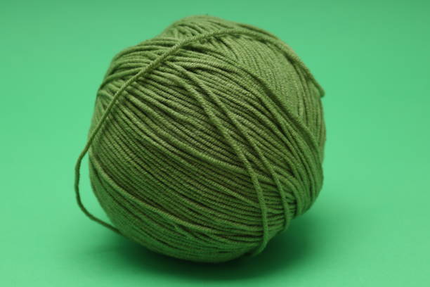 A hand holds a bright pink ball of wool, while several other colorful yarn balls are arranged in the background on a white surface. The colors include shades of green, beige, purple, blue, and grey. The vibrant yarns and neat arrangement create a playful and creative atmosphere, ideal for crafting projects.