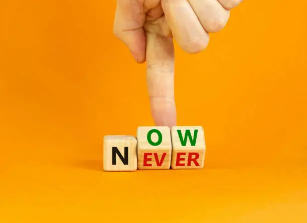 Photo of Now or never symbol. Businessman turns wooden cubes and changes the word 'never' to 'now' or vice versa. Beautiful orange background, copy space. Business and now or never concept.