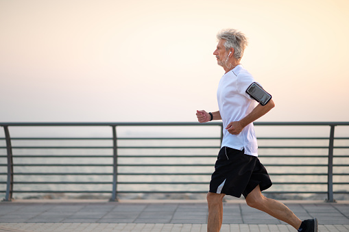 Fit senior Caucasian man jogging on the running track by the seaside outdoors at sunset. Healthy and active lifestyle during retirement. Senior doing sport with a smart watch and a smartphone in a holder on his arm
