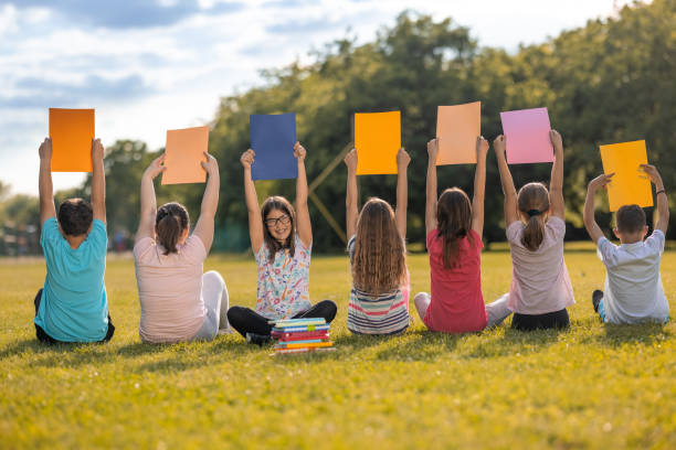 Rear view of elementary students holding a colorful paper while sitting in nature