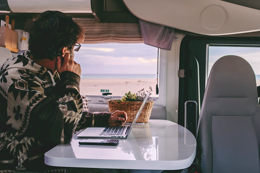 Man working inside camper van sitting at the table and looking outside the window the beautiful beach and ocean in background. Concept of freedom lifestyle and remote online worker people
