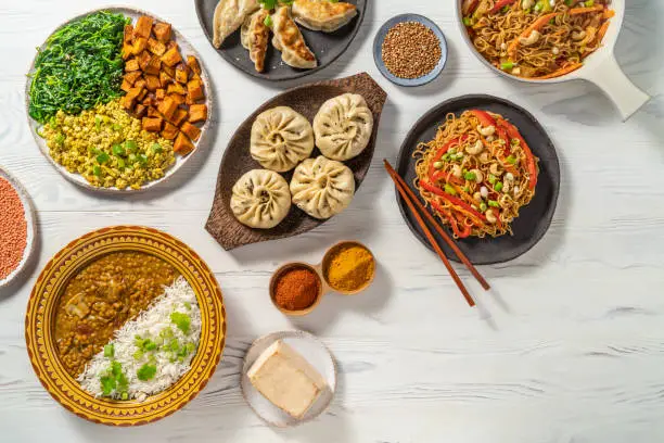 Vegan asian food assortment all plant based recipes as red lentils curry, gyozas, Stir fry Soba noodles, Steamed Panpao and Tofu salad scramble arrangement on white wood background