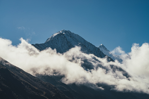 Minimalist view of snow-capped mountain silhouette above thick clouds. Scenic mountain landscape with white-snow sharp peak among dense low clouds in blue sky. Wonderful scenery with snowy pinnacle.