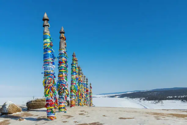 A row of high ritual cult pillars of the Buryats, tied with multi-colored ribbons, on a clear winter day. Sacred Cape Burkhan, Shamanka rock, lake Baikal, Siberia, Russia. Copy space.