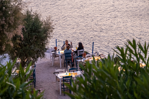 Athens, Greece - September, 29th 2021: View from above behind a tree on a group of white and blue chairs with prepared tables at a sandy pebble beach at dawn. Three ladies sitting at a table next to the shore. Small business restaurant, taverns poster invitation. Beautiful background with copy space.
