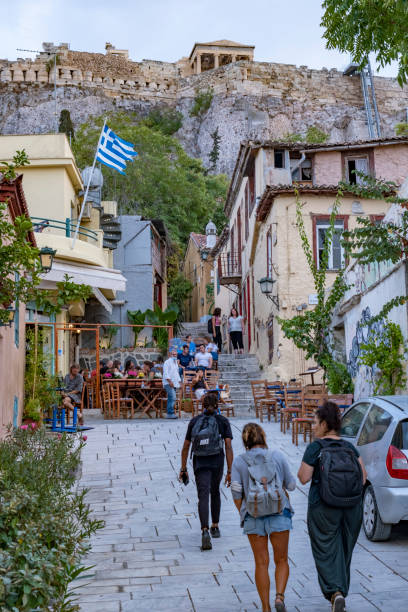 Athens, Greece - September, 28th 2021: Two women walking with backpacks. Beautiful ancient street with green plants. Casual summer clothing. Small group of people sitting outdoors in a street coffee on wooden chairs. On top of the hill the world famous Acropolis Pantheon, a UNESCO World Heritage Site.
