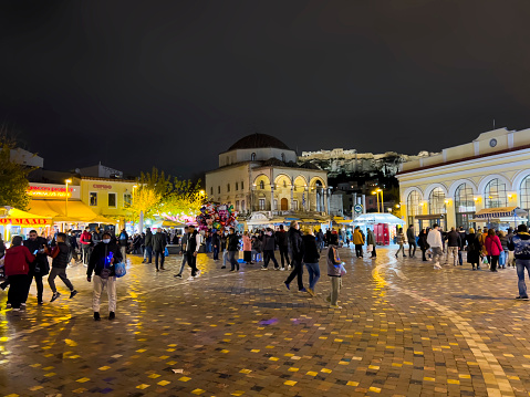 Athens, Greece - December, 30th 2021:  People walking around Monastiraki Square, the flea market neighborhood in the old town of Athens. Photo contains many locals and tourists visiting the square. Stores are open and inviting with bright lights. Dark sky and Acropolis view in background.
