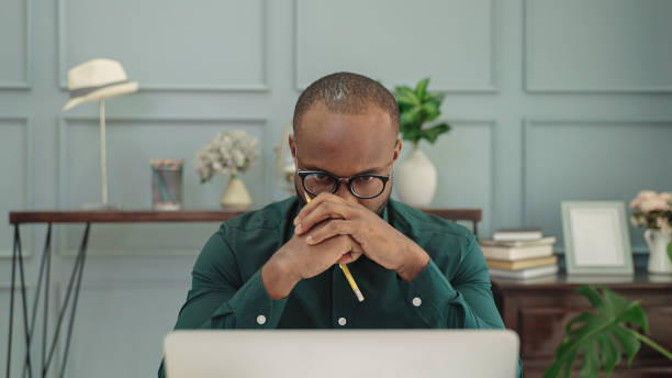 young african American man is thinking in front of a laptop. young african American man is thinking in front of a laptop. in front of stock pictures, royalty-free photos & images