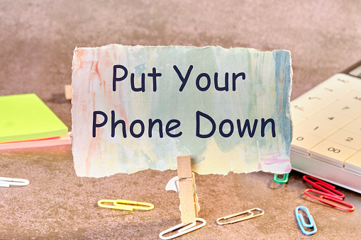 Put Your Phone Down Phrase written on paper next to scattered paper clips, calculator, adhesive paper.