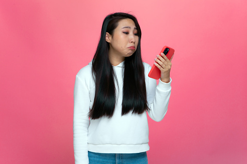 Upset asian young girl looking at phone waiting for call, received refuse, scared of cyberbullying on pink studio background. High quality photo