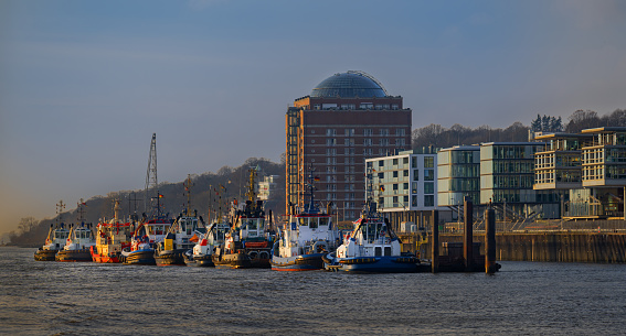 Tug boats moored at a pier. Tug brigde with moored tug boats. Tugboats moored at bollards by the pier New tugboat bridge in Neumühlen Hamburg. Tugboats docked on the banks of the Elbe river.
