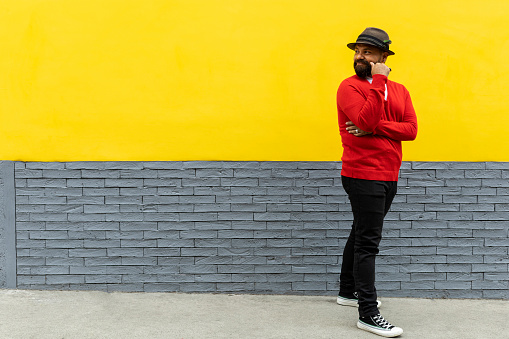 Adult Latino male (39) with beard, standing and looking behind him with a gesture of searching for something. Wearing hat, red sweater and white flannel. Yellow and gray wall background. Space to copy. Body language concept