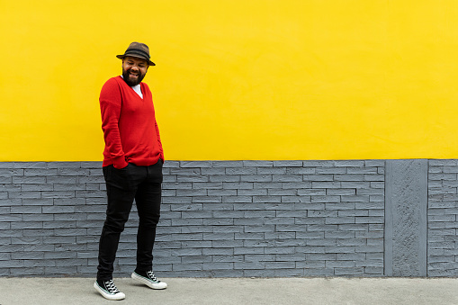 Adult Latino male (39) with beard, standing with his hands in his trouser pockets, making a cheerful gesture. He wears a hat, red sweater and white flannel. Yellow and gray wall background. Space to copy. Body language concept
