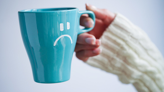 Blue Monday concept. Close-up of a female hand in a white sweater holds a blue cup with a sad smiley as a symbol of the most depressive day of the year on a white background