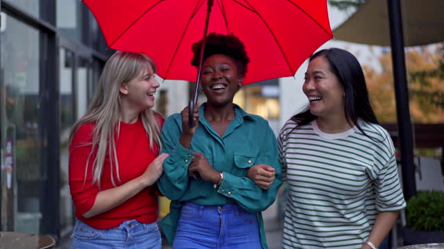 Portrait of an cheerful urban city multiracial female friends walking under umbrella during rainy day in the city