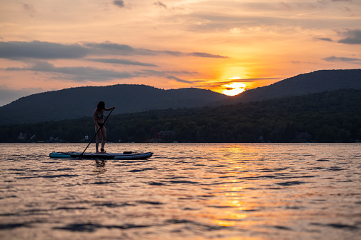 Woman Paddleboarding on the Lake at Sunset in Summer. It is a beautiful summer evening, the lake is calm and the sky is colourful, romantic sky. She is standing on her paddleboard and looking at the sunset. Lac Saint-Joseph, Quebec, Canada.