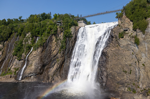 Montmorency Falls in Summer, Quebec City, Canada. It is a beautiful sunny summer day and there is a rainbow. Tourists are walking on the suspension bridge.