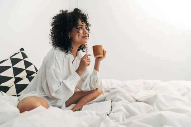 Young mixed race woman sitting on bed having coffee and writing on her journal. Copy space