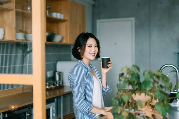 Photo of Relaxed young Asian woman enjoying a cup of morning coffee by the kitchen counter at home, day dreaming while looking away