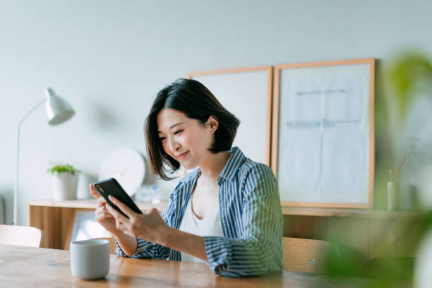 bella giovane donna asiatica sorridente che gestisce il pagamento delle bollette finanziarie con smartphone e carta di credito nel soggiorno di casa. la tecnologia rende la vita molto più facile - smart phone asian ethnicity mobile phone women foto e immagini stock