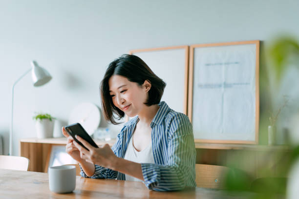 Beautiful smiling young Asian woman managing financial bill payment with smartphone and credit card in the living room at home. Technology makes life so much easier stock photo