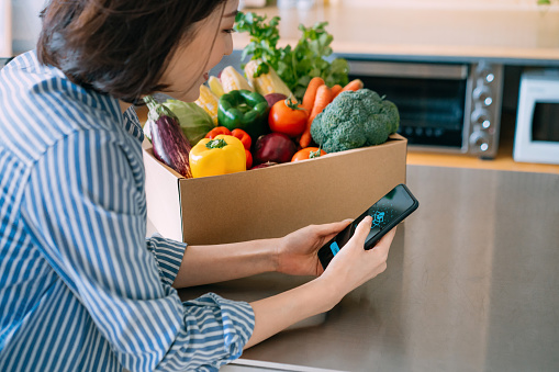 Smiling young Asian woman doing home delivery grocery shopping online with mobile app device on smartphone at home, with a box of colourful and fresh organic vegetables and fruits on the table
