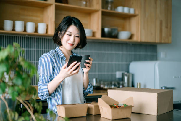 istock Beautiful smiling young Asian woman standing by the kitchen counter, using smartphone while enjoying assorted home delivery takeaway food at home. Eating at home concept 1364894347