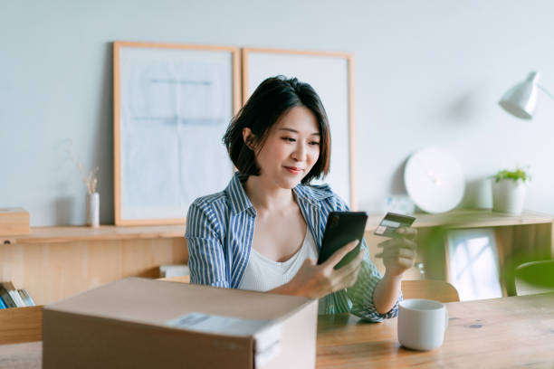 jeune femme asiatique gaie faisant des achats en ligne avec le smartphone et faisant le paiement mobile avec la carte de crédit à portée de main, recevant un colis par service de livraison à domicile. la technologie rend la vie tellement plus facile - qr code marketing mobile phone coding photos et images de collection