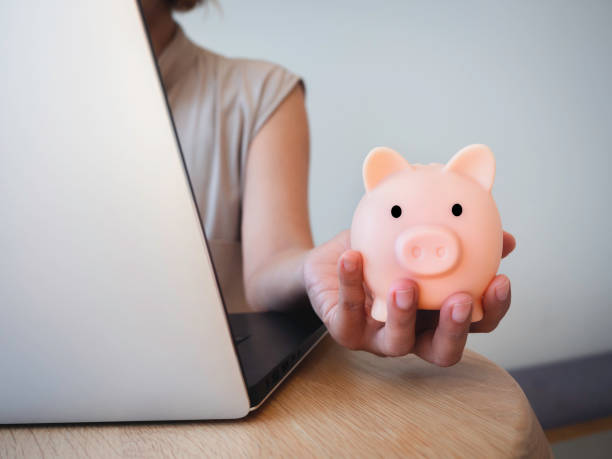 istock Money box on hand. Hand holding the pink piggy bank while using laptop computer on desk. Saving money wealth and financial concept, Business, online banking, finance, investment, financial planning. 1364903564